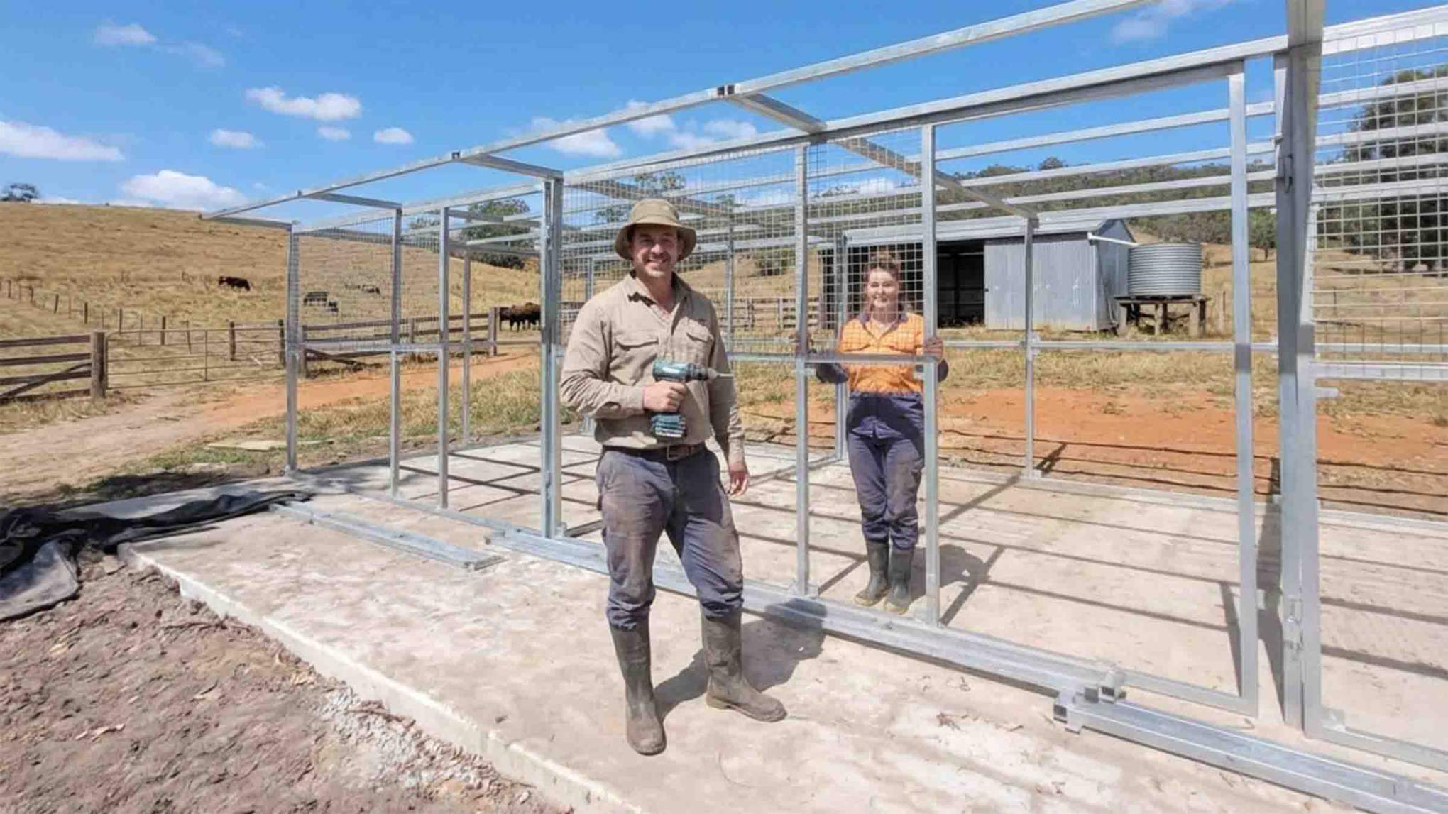 Two people assembling a modular steel horse stable frame on a rural property, using pre-welded mesh panels and tools on a concrete base.