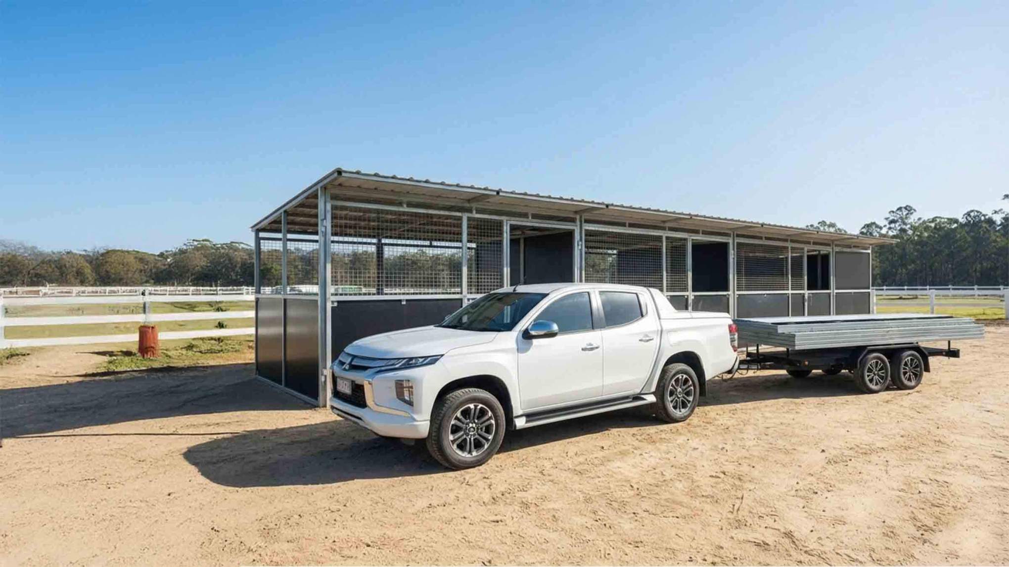 White utility vehicle and trailer parked beside a 4-bay modular steel horse stable with mesh panels and flat roof on a rural property.