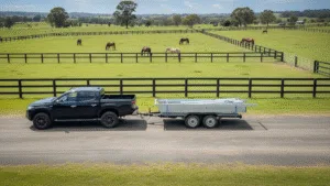 Ute towing trailer with flat pack stable on country road, horses in paddock.