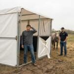 Two men inspect a damaged portable stable made from thin metal and fabric panels, showing alignment and material issues typical of cheap imported kits.